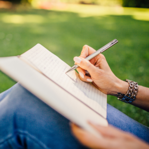 Person writing in a notebook with a pen while sitting on grass in a park.