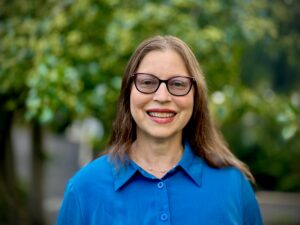 Gabrielle Kaplan-mayer is a woman with long brown hair and glasses, smiling, wearing a blue shirt, standing outdoors with greenery behind her.