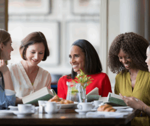 Four women sit at a table, smiling and reading books together in a cozy, well-lit café.