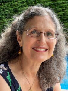 Smiling woman with gray curly hair and glasses, wearing earrings, outdoors near greenery and a blue background.