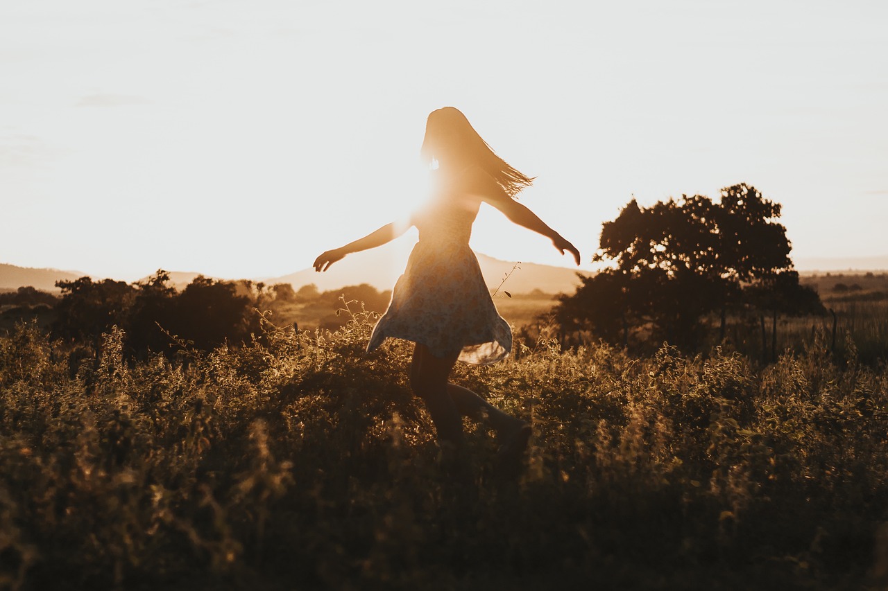 shot of the silhouette of a person standing in a field with flowing hair wearing a dress with outstretched arms