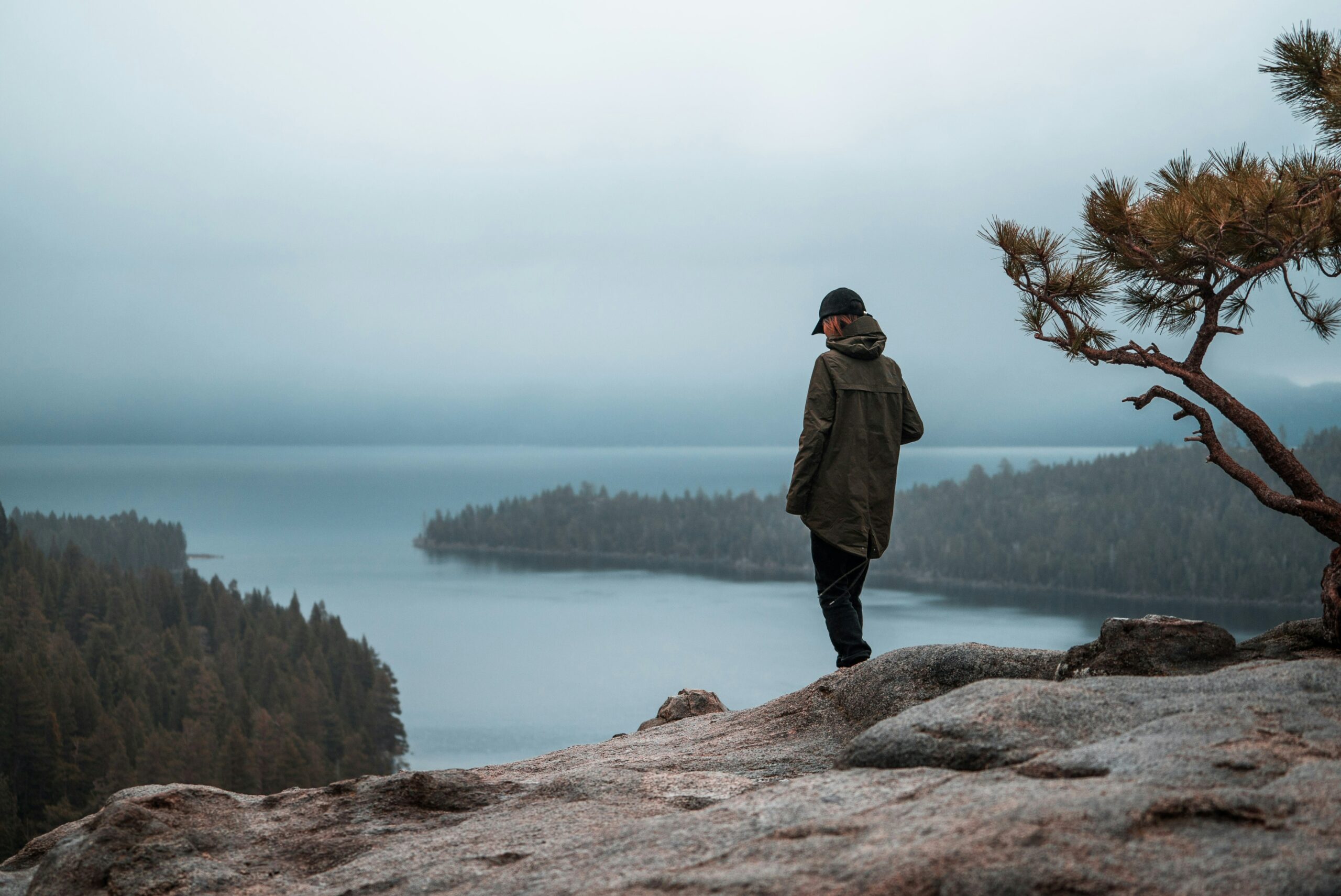 a person in a jacket and hat is standing at the edge of a cliff overlooking Emerald Bay on a foggy day