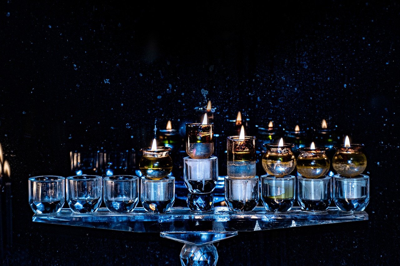 A chanukiah made of oil candles sits on a reflective surface against a dark background.