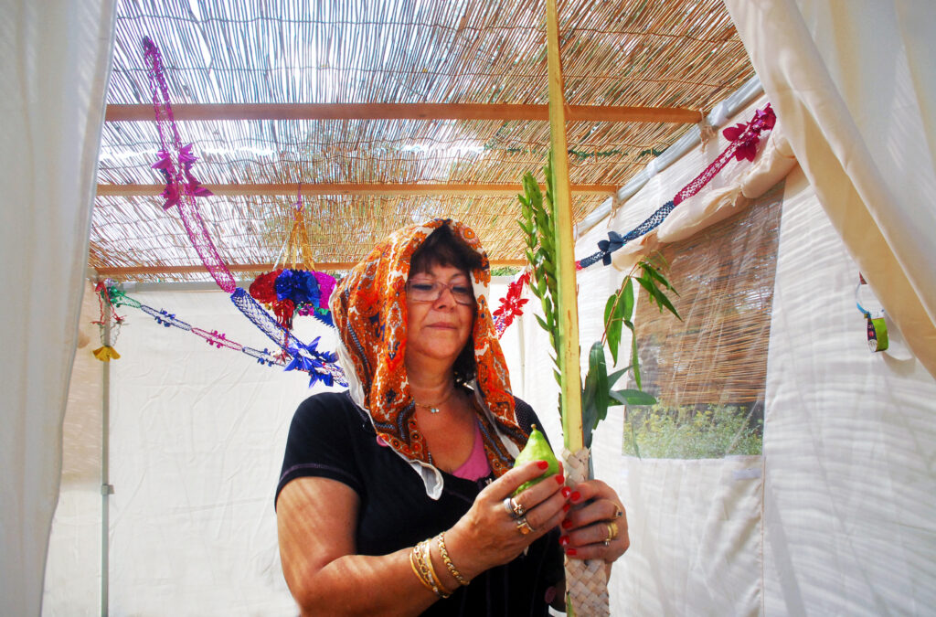 woman with headscarf draped on her head holding lulav and etrog inside a sukkah