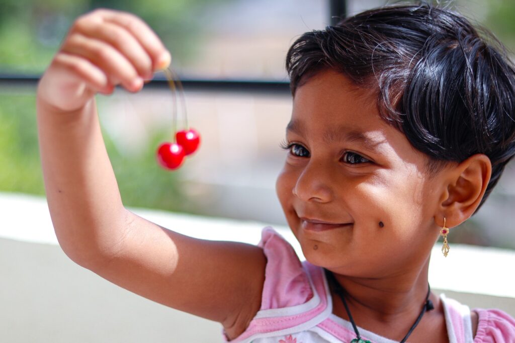 a child with short hair and earrings smiles, holding up and looking at a couple of cherries