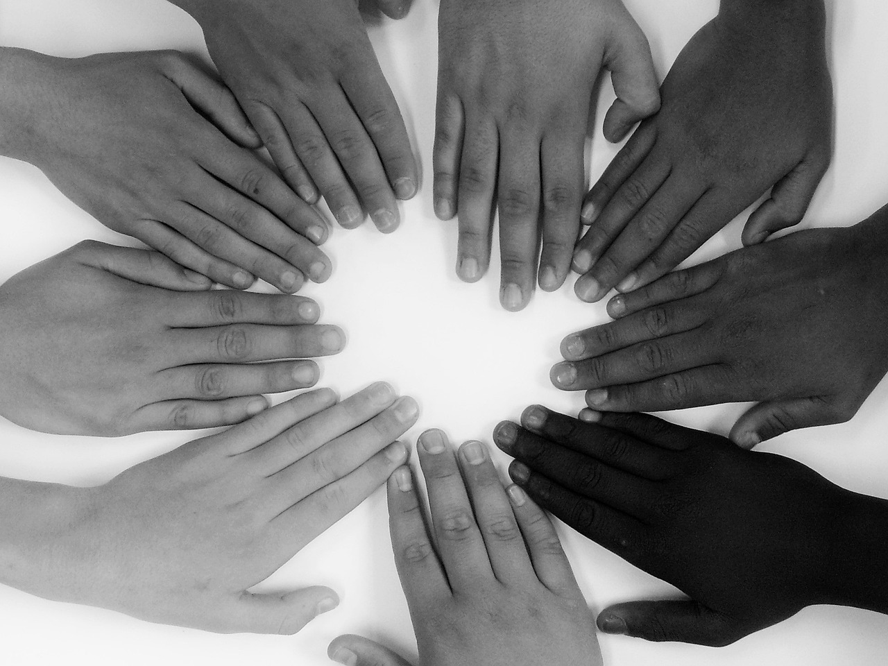 black and white photo of hands of different skin tones lying flat on a surface and arranged in a circle with fingers pointing toward the middle