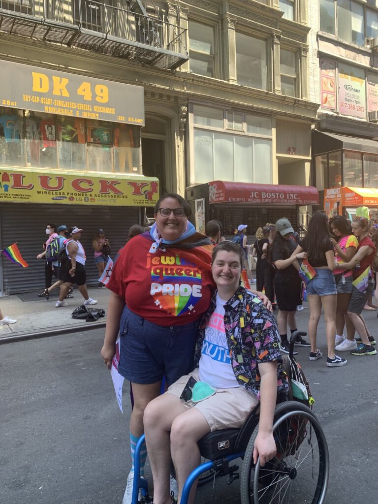 Two people at a pride parade, one standing with a rainbow flag, the other in a wheelchair, both smiling.