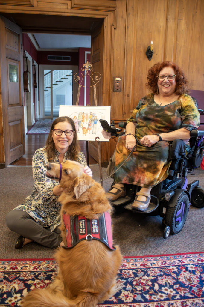 Two women, one in a wheelchair, smiling at a service dog in a room with wooden walls and a colorful carpet.