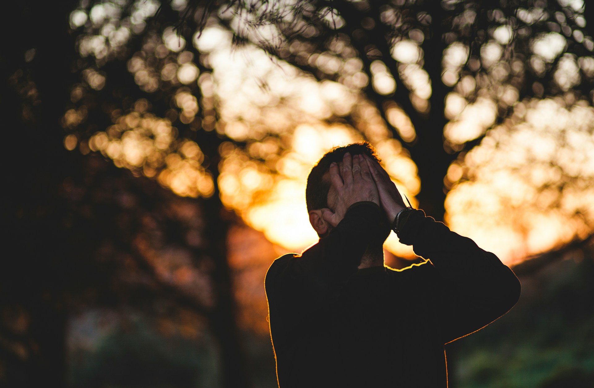 a person stands outside near trees covering their face with their hands