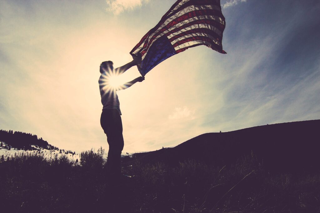 person holding large american flag as it waves in the wind against a shining sunlit sky