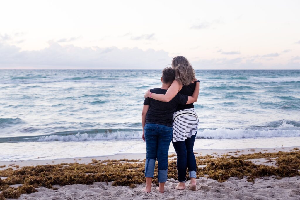 mother and teen hug facing the water on the beach