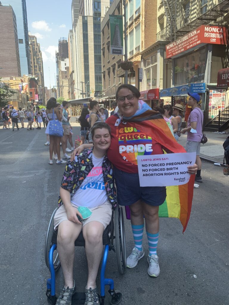 Two people at a Pride event; one in a wheelchair, the other draped in a rainbow flag with a rights sign.
