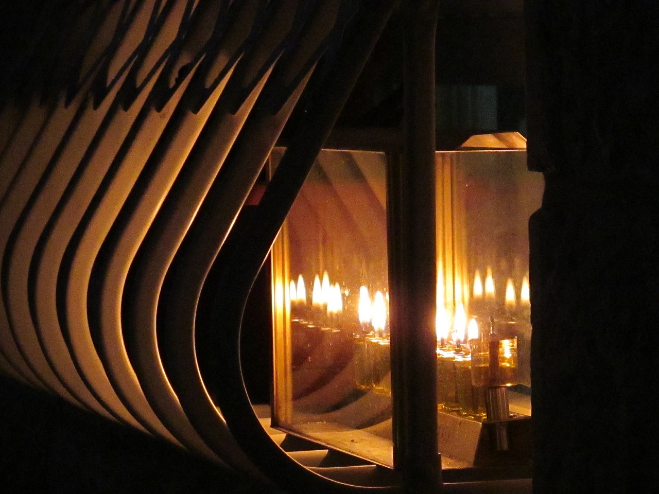 A close-up of candles lit in a reflective menorah, casting warm light and shadows through a geometric pattern.