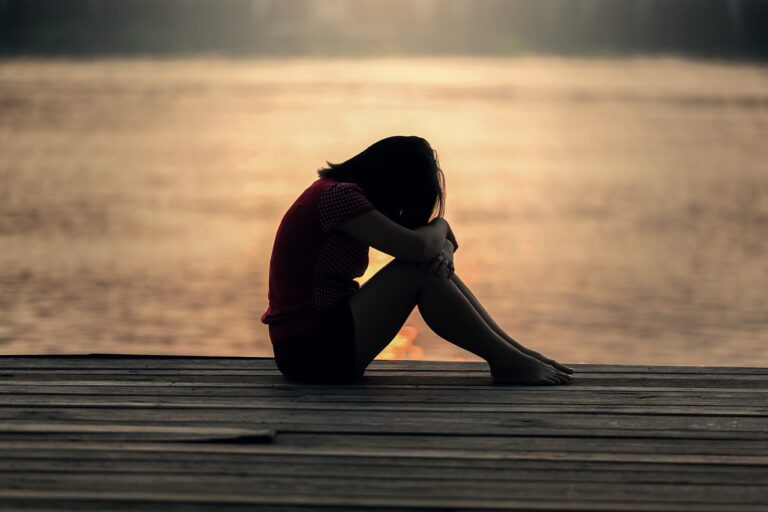 the silhouette of a child with medium length hair is sitting on the ground with their head on their needs, in the background is a body of water