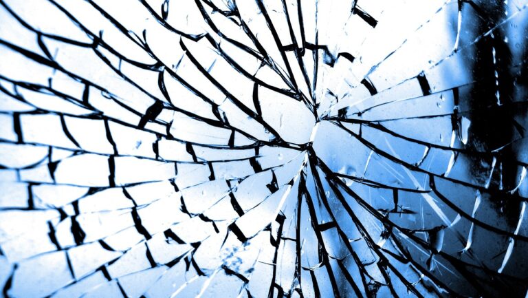 Close-up of shattered glass with sharp cracks radiating from a central point, tinted in blue tones.