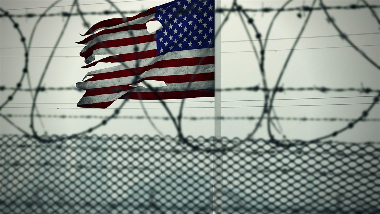 a barbed wire fence stands in front of a torn American flag waving against a gray sky