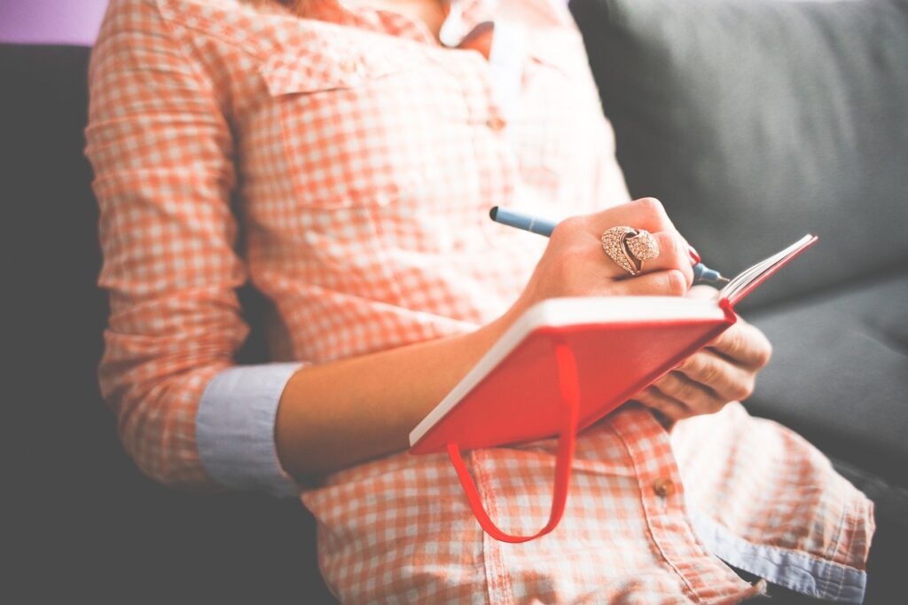 A person in an orange checked shirt wearing a ring writes in a red journal