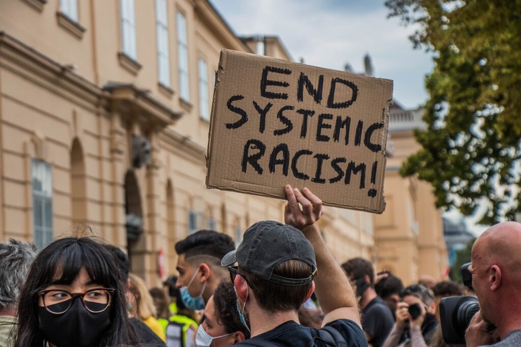 a protest, several people are wearing masks and someone is holding a cardboard sign that says "END SYSTEMIC RACISM"