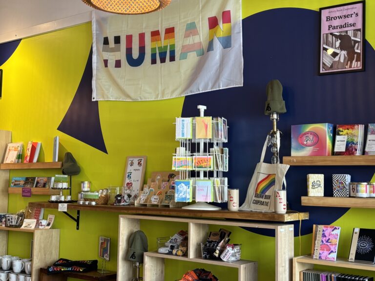 Colorful shop display with pride-themed items, books, and a HUMAN banner on a yellow and blue wall.