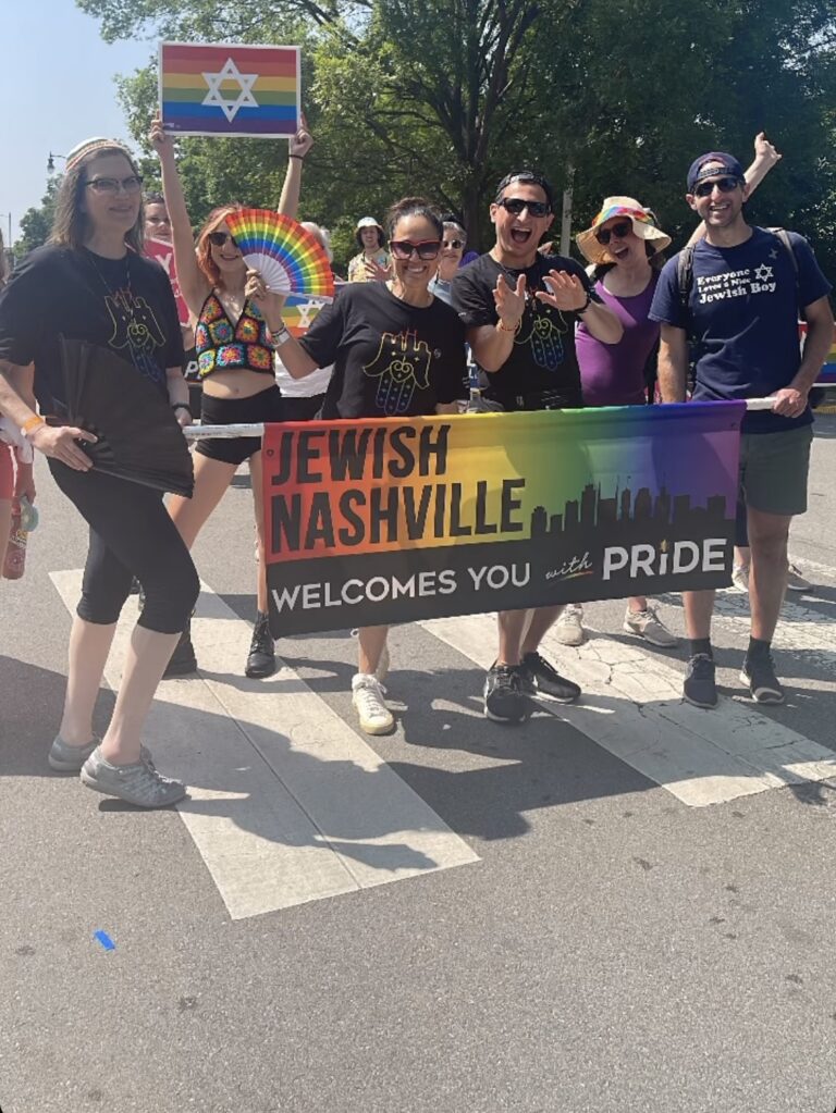 A group holds a Jewish Nashville Welcomes You with Pride banner at a Pride parade on a sunny day.