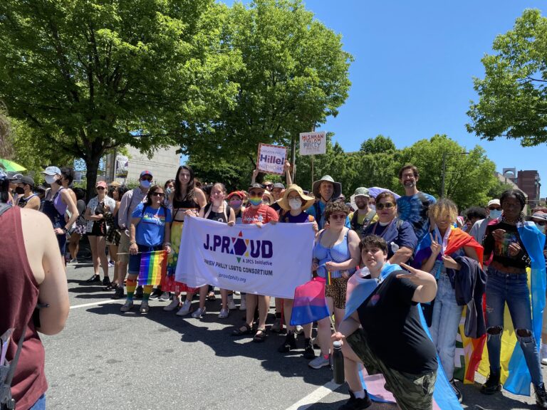 A group of people at a Pride parade hold a “J.Proud” banner, surrounded by rainbow flags and signs.