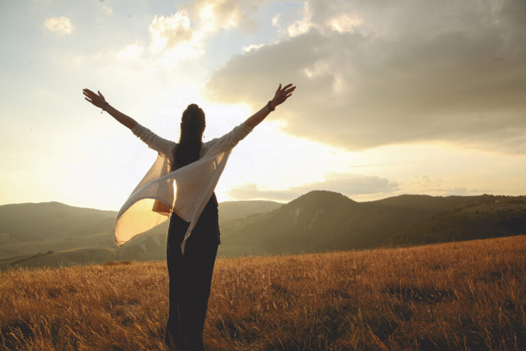 Person standing in a field at sunset, arms raised, enjoying the open landscape and warm light.