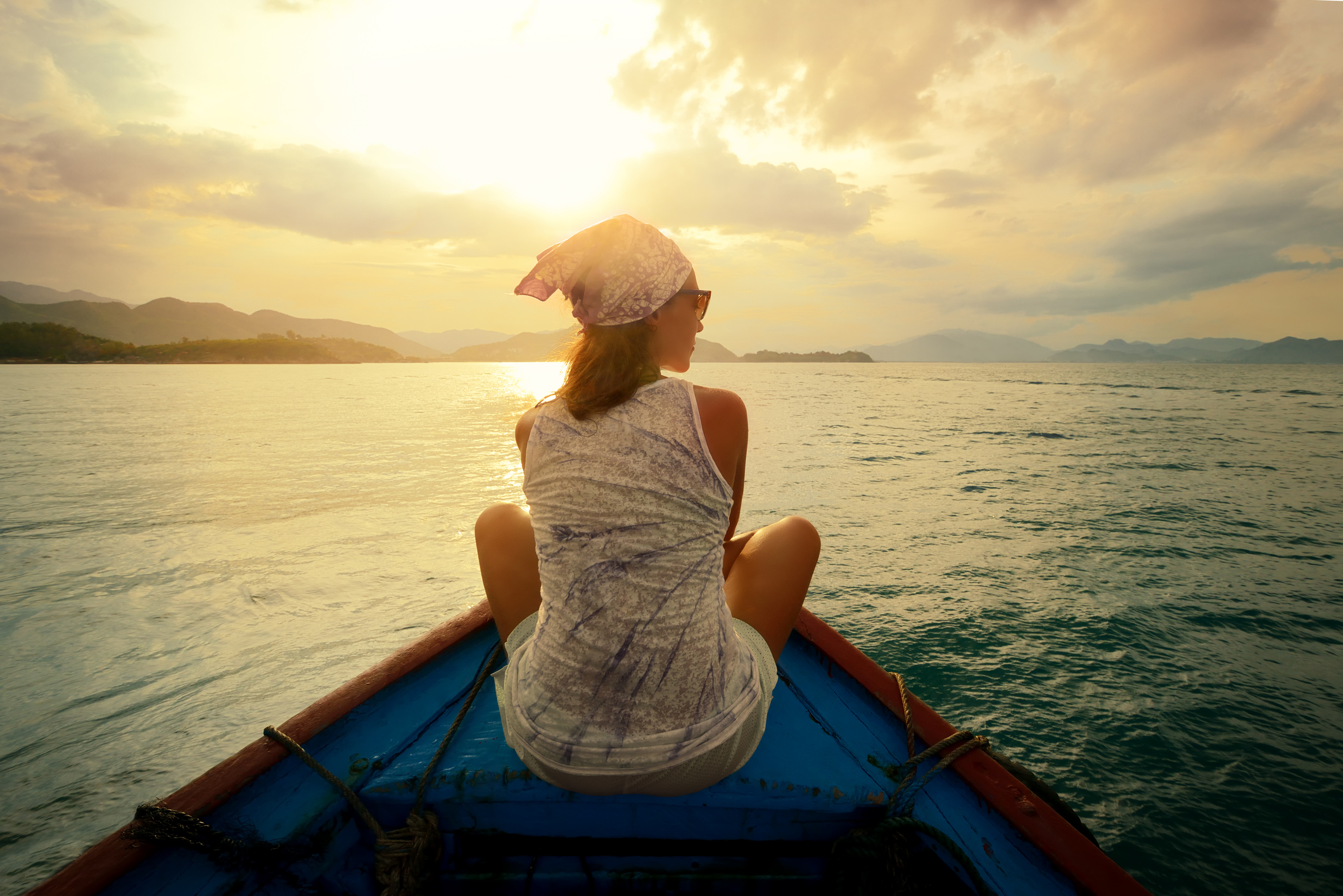 A person sits at the front of a boat, gazing at a sunset over a calm sea, with hills in the distance.