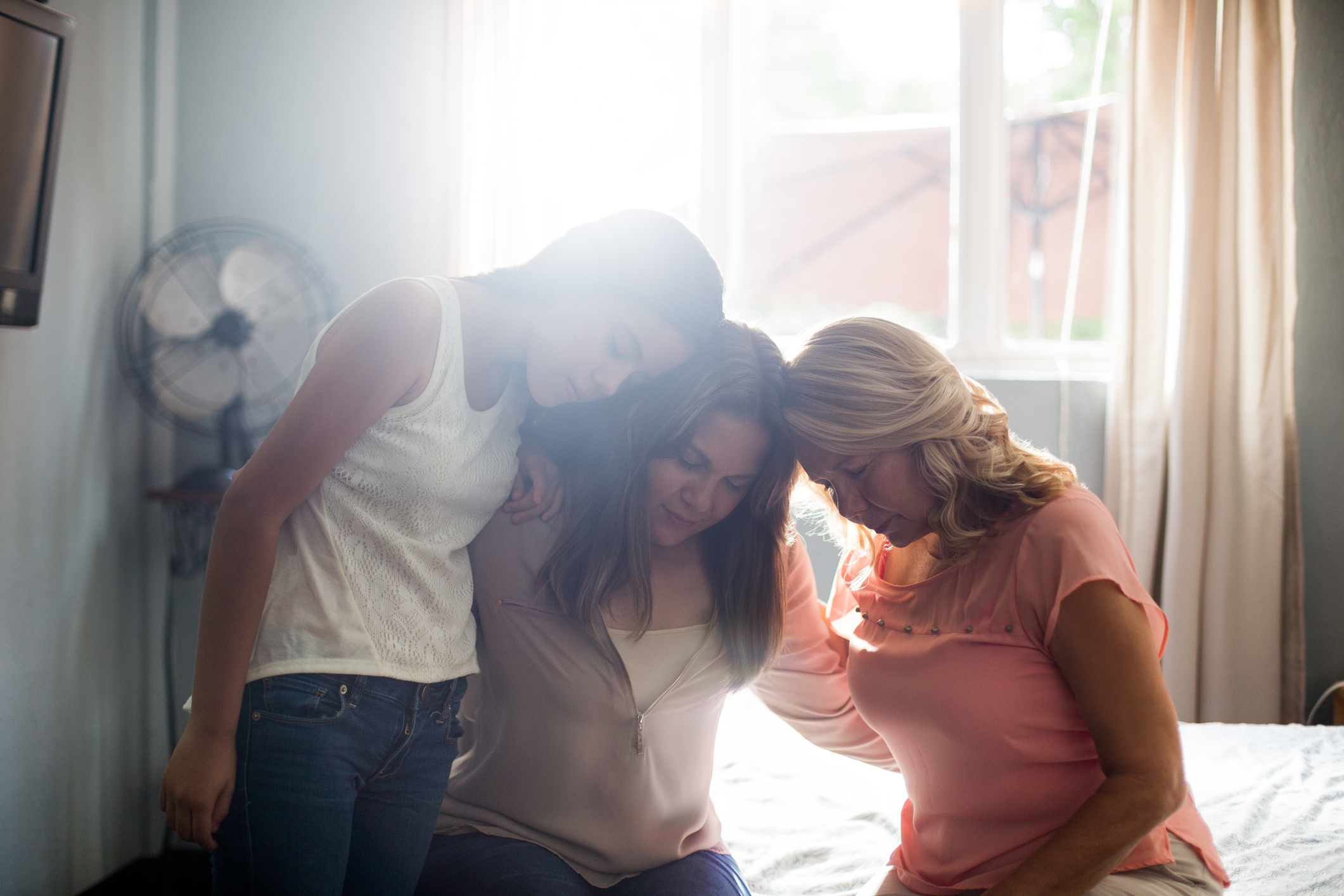 Three women sit close together on a bed, comforting each other with their heads touching, in a softly lit room.