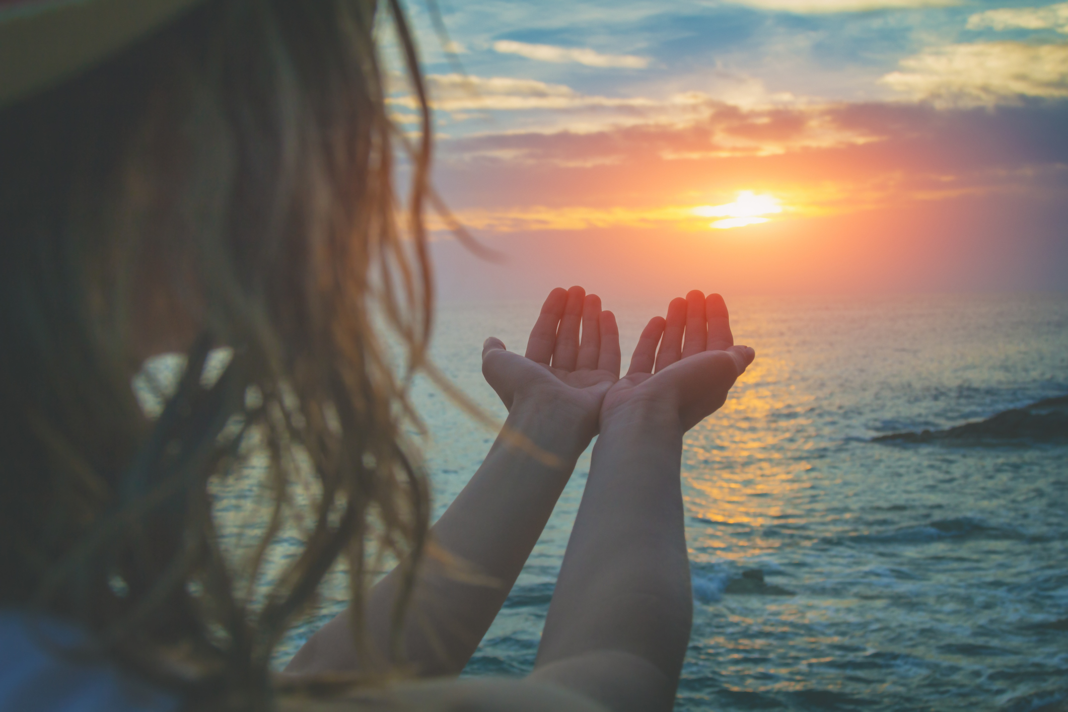 Person holding hands up toward ocean sunset, with warm colors in the sky and hair blowing gently.