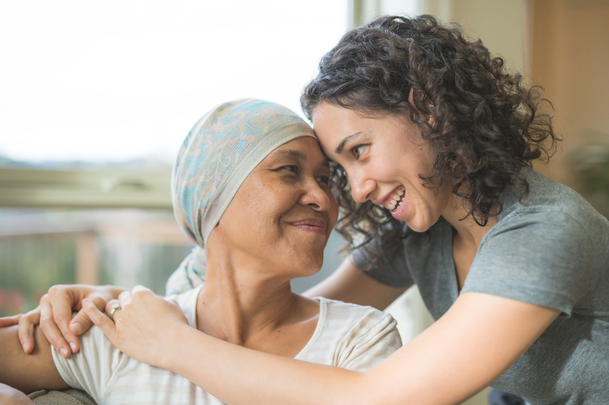 A woman in a headscarf is embraced by a smiling young woman at home.