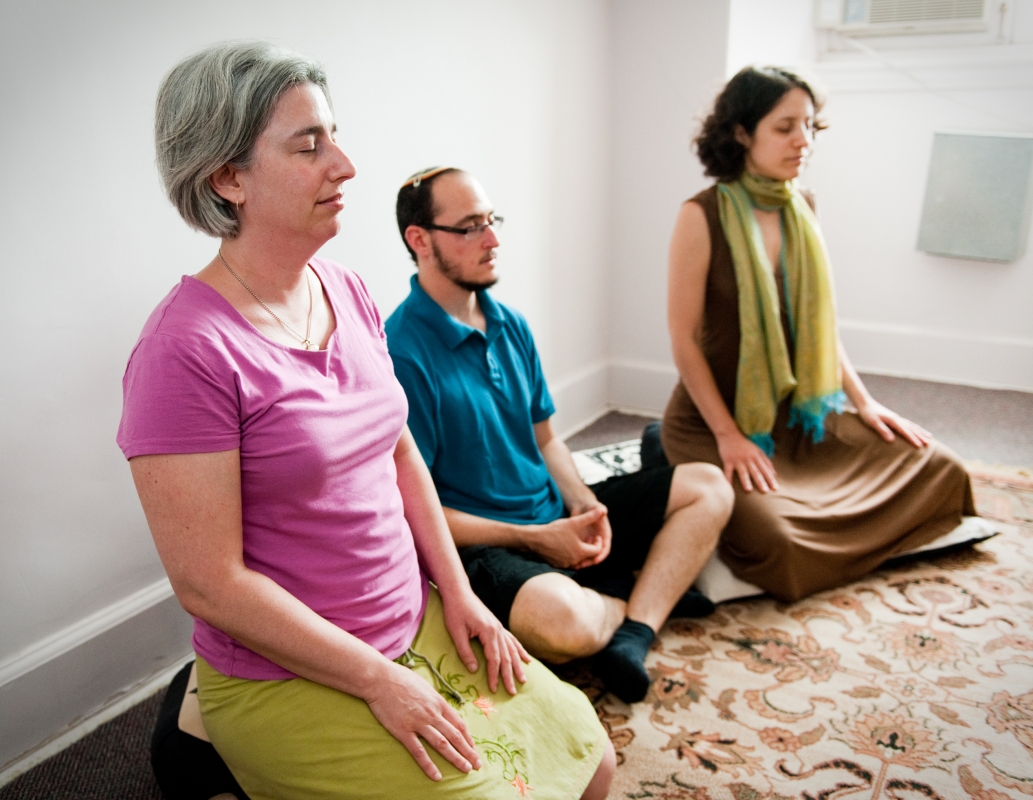 Three people sitting cross-legged on the floor, meditating in a serene room with minimal decor.