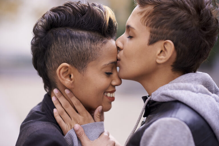 Two women with short hair embracing, one kissing the other's forehead outdoors, sharing a tender moment.