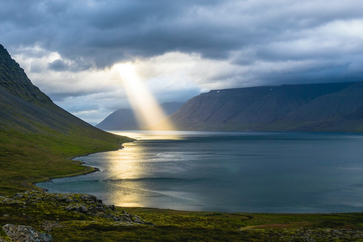 Sunbeam breaks through clouds, illuminating a serene fjord surrounded by mountains.