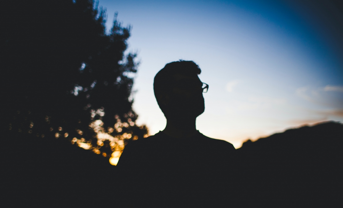 Silhouette of a person with glasses against a sunset sky, surrounded by dark trees.