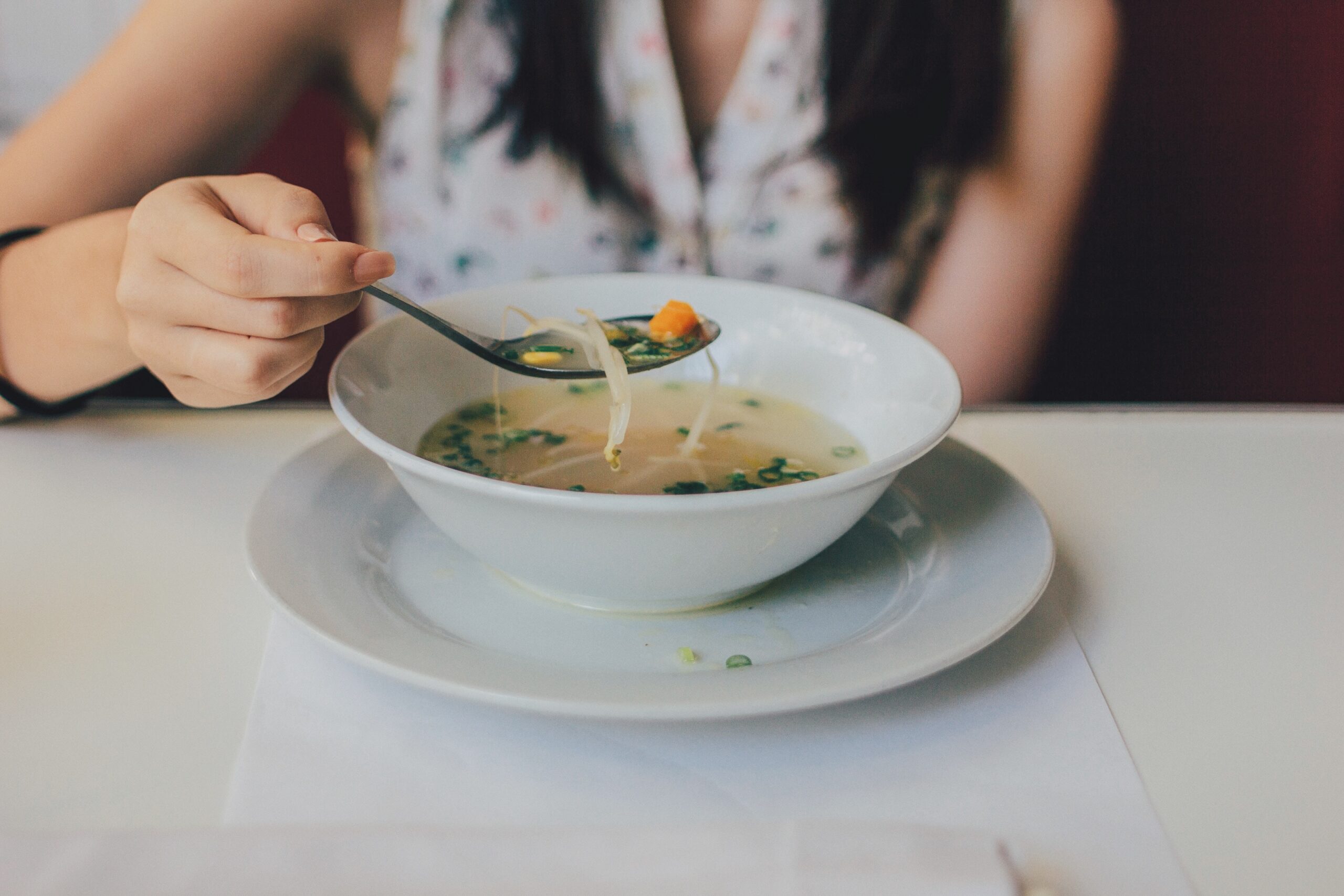 A person is holding a spoon above a bowl of noodle soup on a white table.