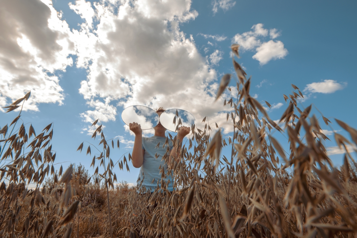 Person in field holding two mirrors reflecting clouds, surrounded by tall grasses under a partly cloudy sky.