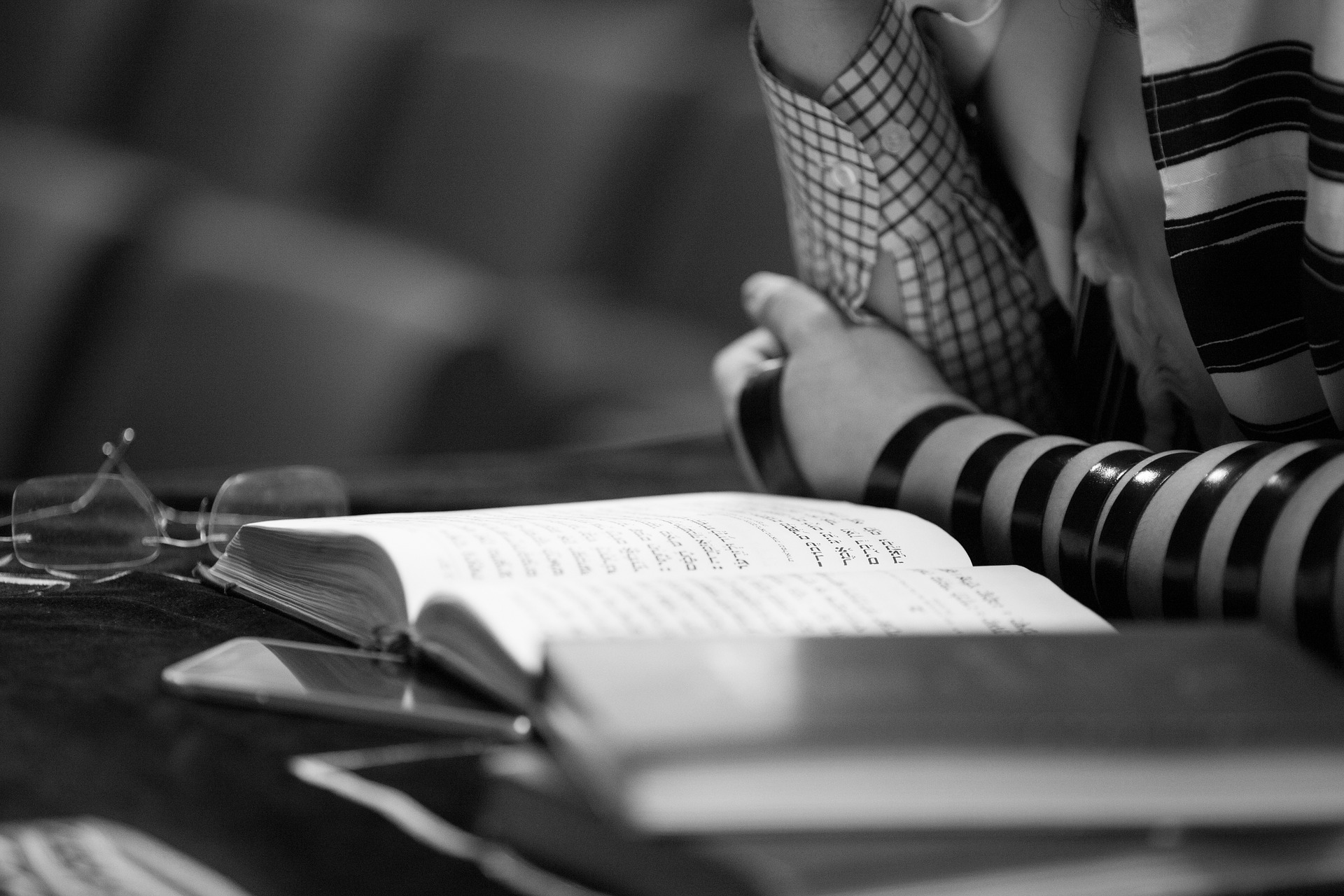 Person praying with Hebrew prayer book and tallit, seated at a table with glasses and additional books, in grayscale.