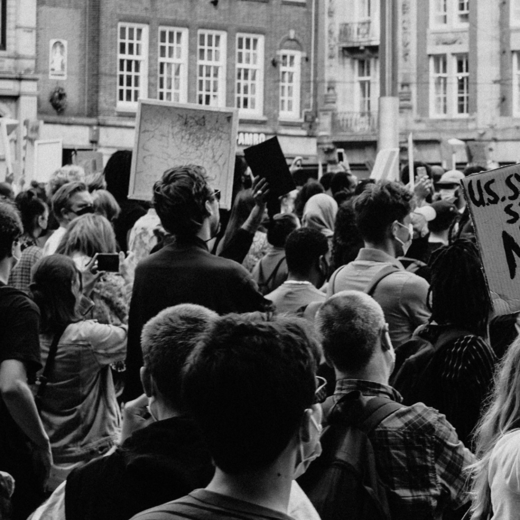 Crowd at a protest, holding signs. Urban street setting with old buildings in the background.