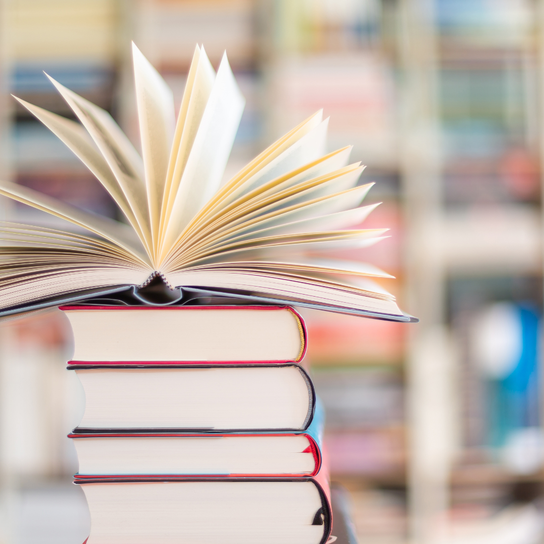 An open book on top of a stack of books with blurred shelves in the background.