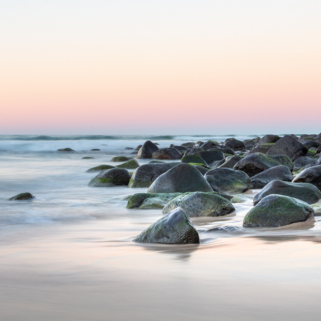 Sunset over a rocky shoreline, with stones covered in moss and calm waves lapping the beach.