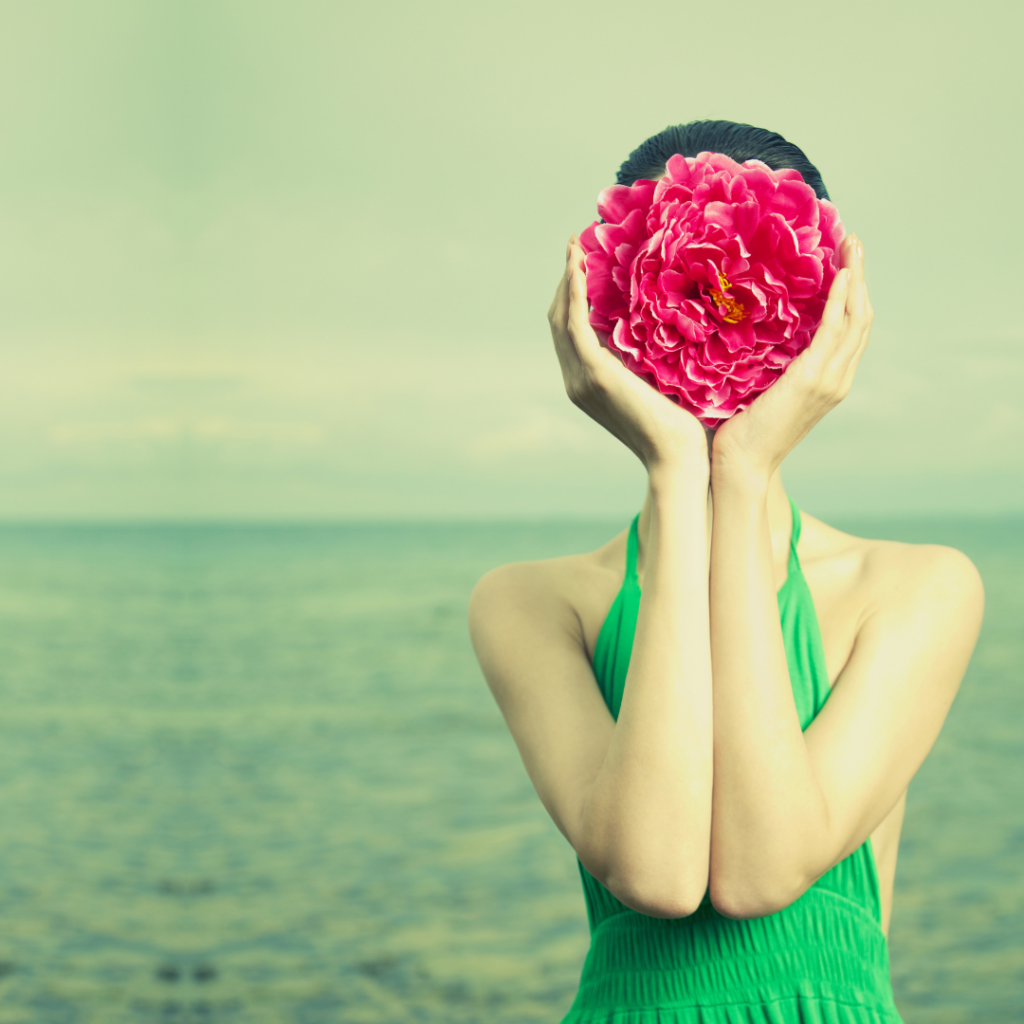 white woman standing in front of ocean holding fuschia flowers in front of her face