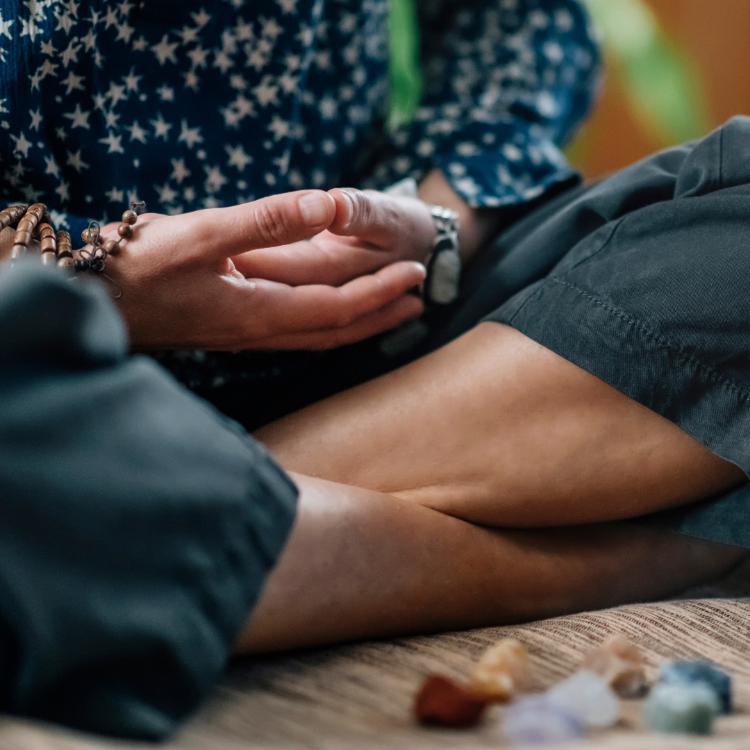 Person sitting cross-legged, holding mala beads, surrounded by crystals.