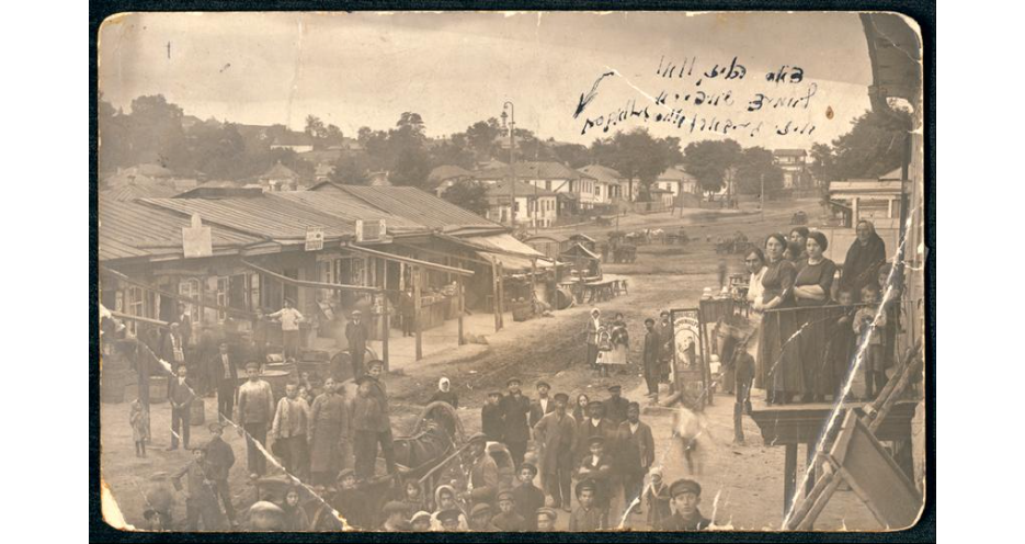 A sepia-toned old photograph of a bustling village street with people and buildings.