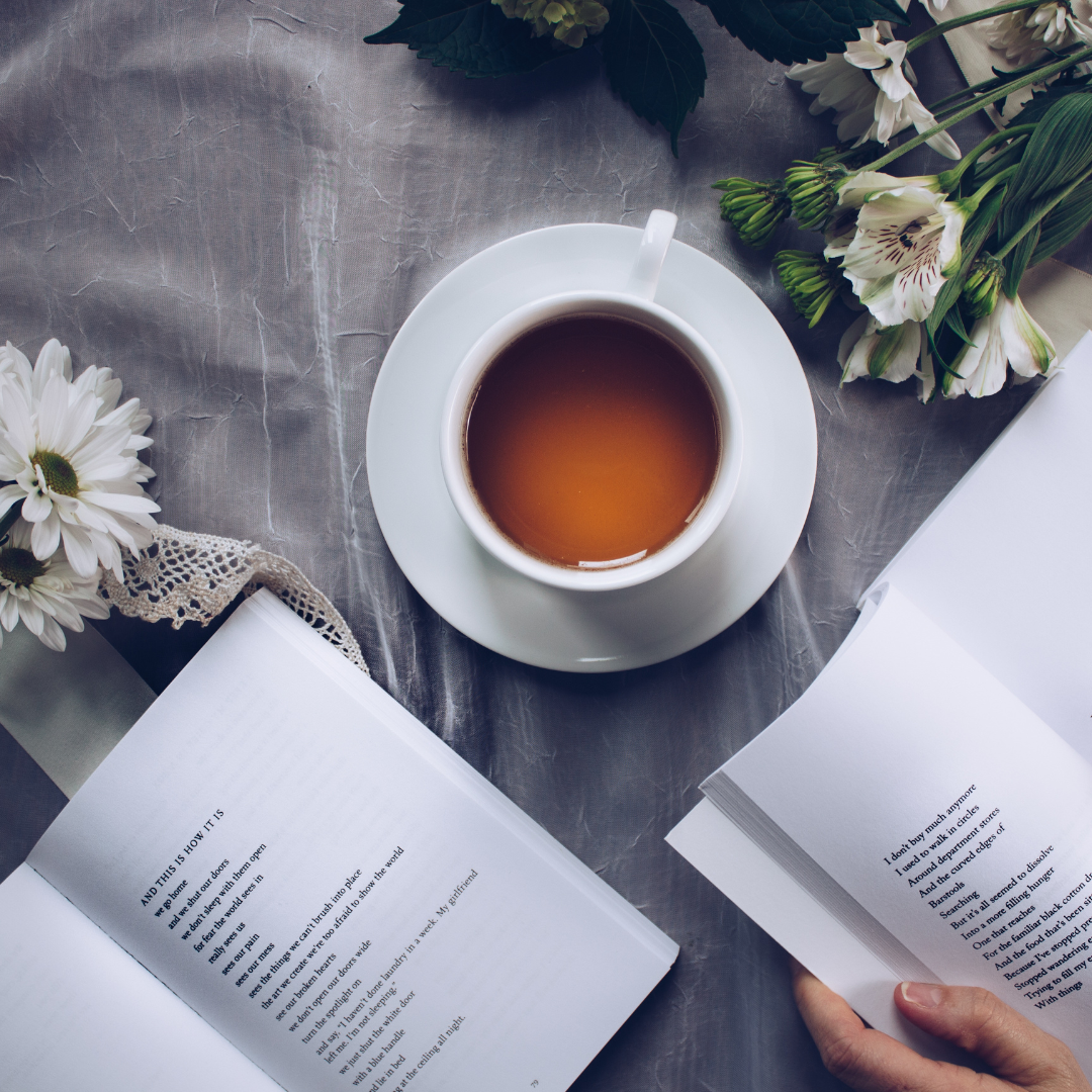 A cup of tea on a saucer with open books and flowers on a gray surface.