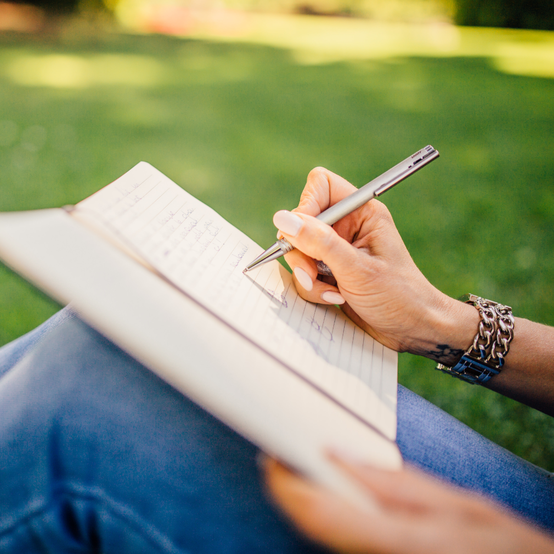 Person writing in a notebook while sitting on grass outside.