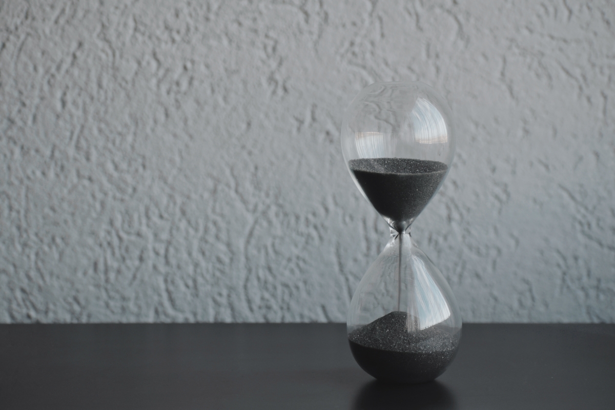 An hourglass with black sand sitting on a dark surface against a textured gray wall.