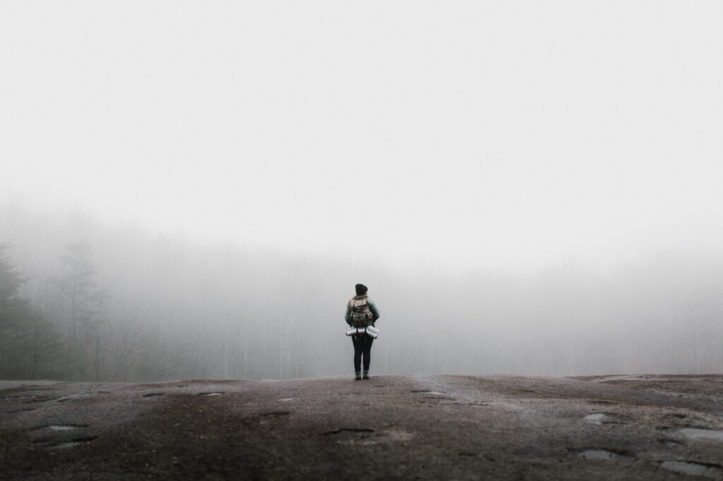 person with backpack on standing outdoors looking out onto a misty sky with blurry landscape in the distance