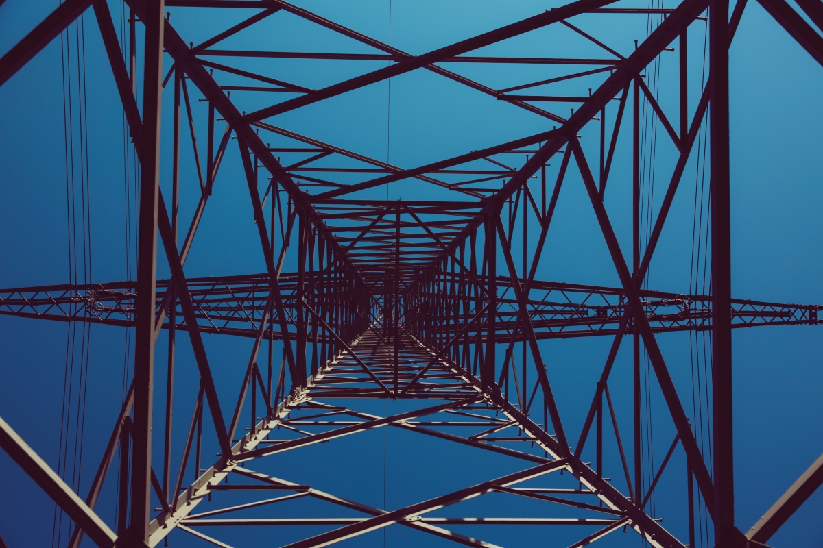 View from below of a power transmission tower against a clear blue sky, showcasing its geometric metal structure.