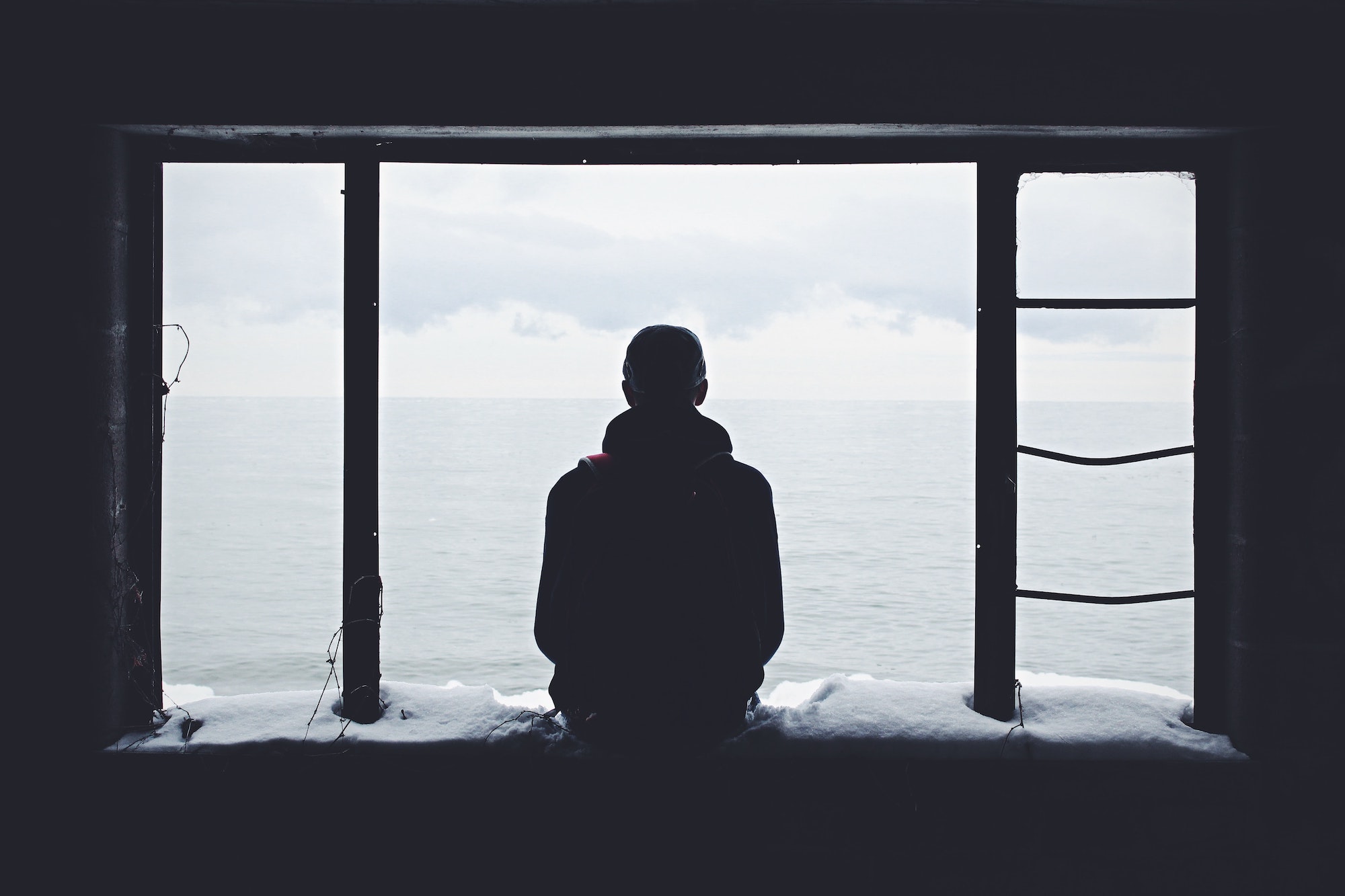black and white photo of man in silhouette sitting at the ledge of open window looking out at cloudy sky