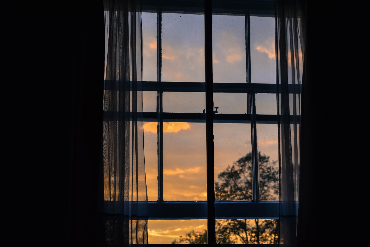 Sunset view through a window with sheer curtains, silhouetted trees outside.